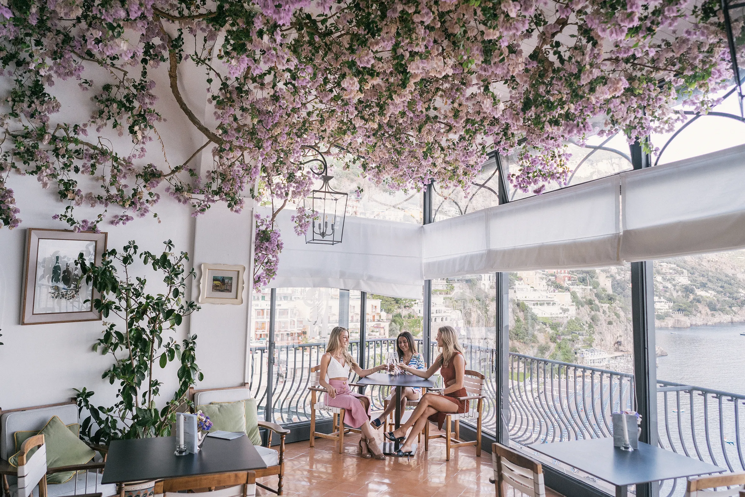 Group of women celebrating with wine glasses on a balcony overlooking Positano and the Mediterranean sea.