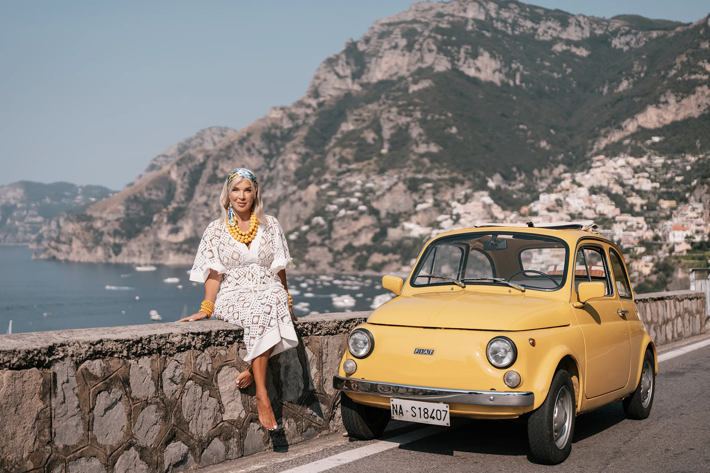 Woman in white dress sitting by a vintage yellow Fiat 500 overlooking Positano on the Amalfi Coast, Italy