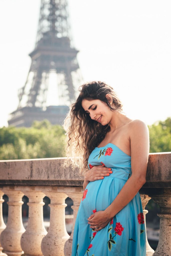 Pregnant woman smiling near the Eiffel Tower in Paris Pregnant woman in a blue floral dress standing by a stone balustrade with the Eiffel Tower in Paris in the background