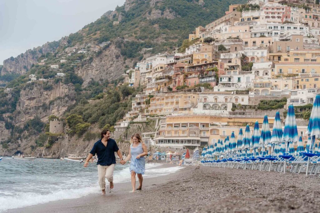 Couple walking along the beach in Positano Couple holding hands while walking along the beach in Positano with cascading cliffside buildings behind them