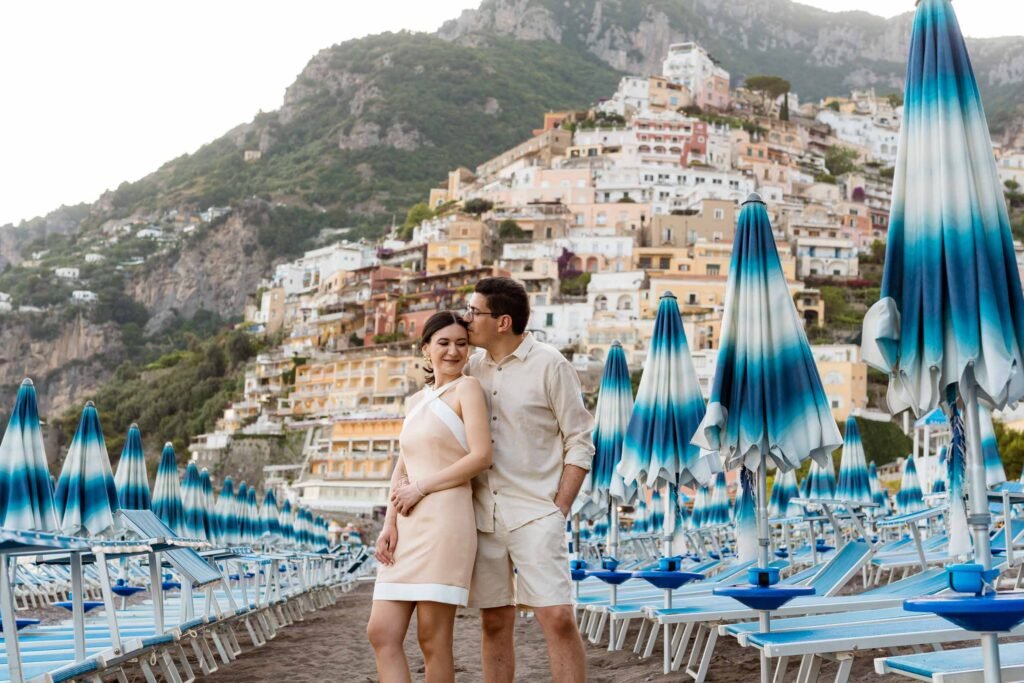 Couple on the beach in Positano with scenic coastal views Couple posing on the beach in Positano with blue umbrellas and cliffside buildings