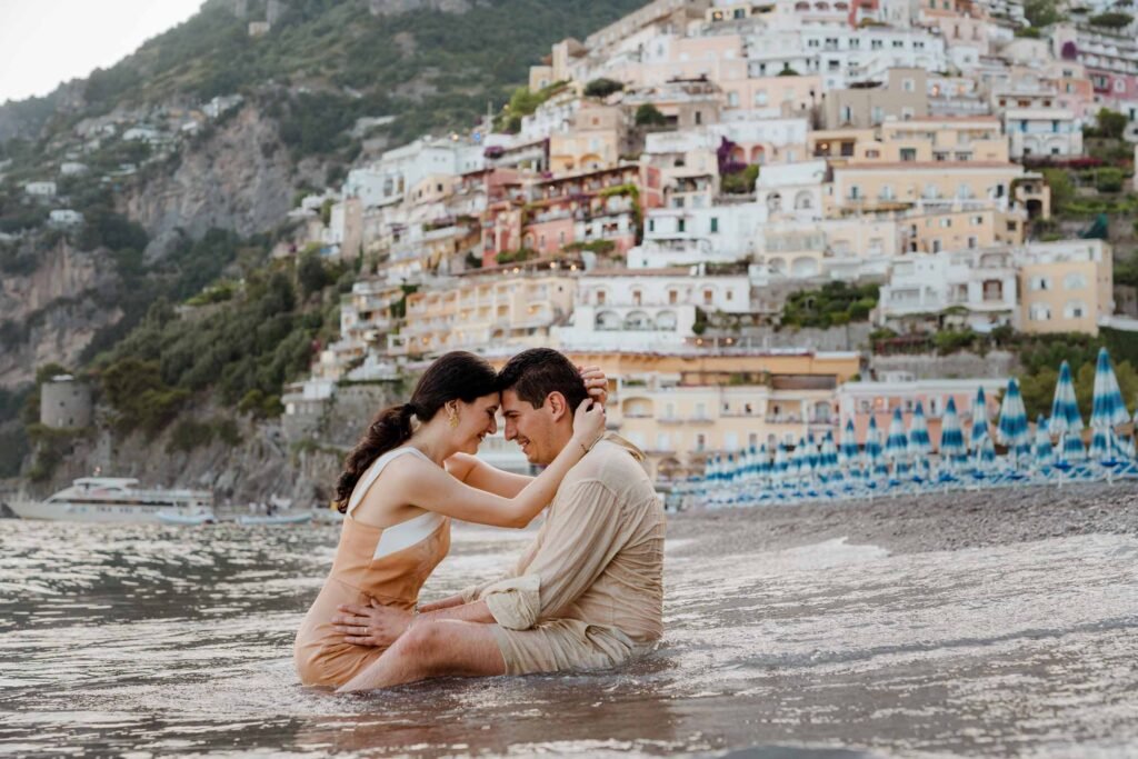 Couple sitting in the shallow water at Positano beach Couple sitting in the shallow water on the beach in Positano with cascading cliffside buildings behind them