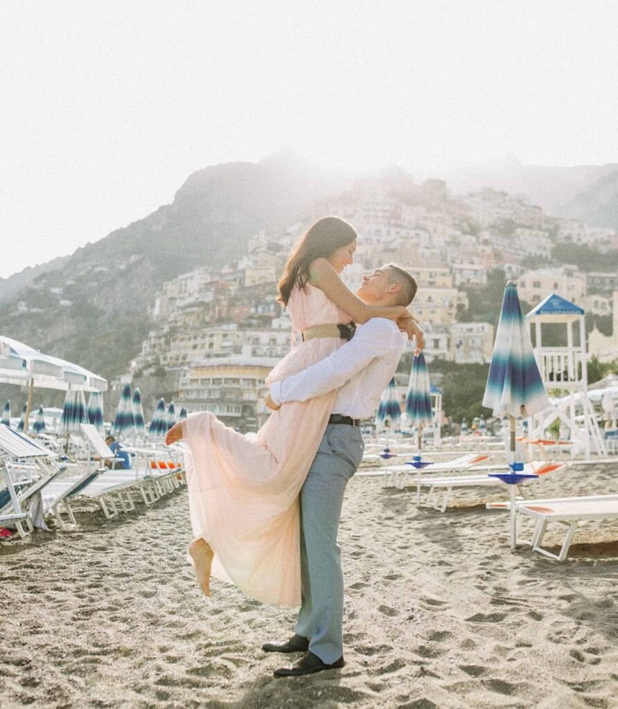 Couple embracing on the beach in Positano Man lifting a woman in a long dress on the beach in Positano with a coastal town background