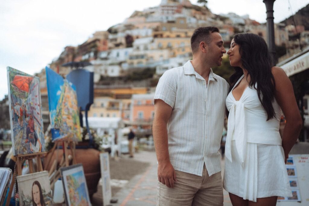 Couple walking along the coast in Positano Couple leaning in close to kiss on a walkway in Positano with paintings and colorful buildings in the background