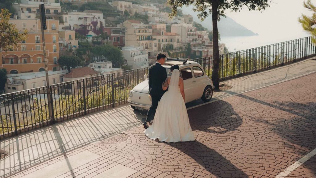 Bride and groom standing by vintage car in Positano Bride in a white gown and groom in a suit standing next to a classic white car overlooking Positano