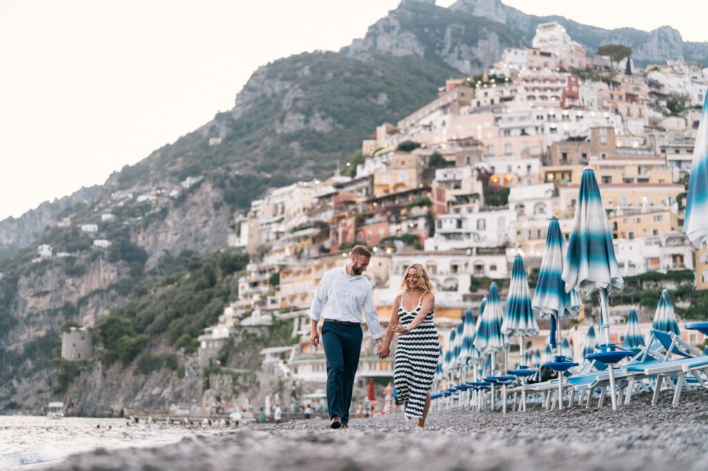 Portfolio Photo Couple holding hands while walking on the pebble beach in Positano with cliffside buildings behind them