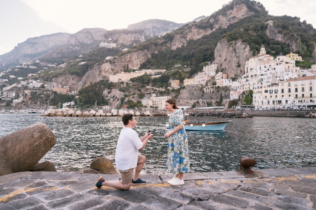 Portfolio Photo Man kneeling to propose to a woman by the harbor in Amalfi with coastal mountains in the background