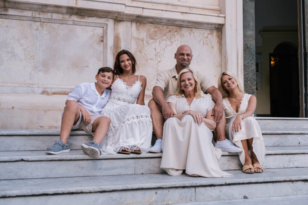 pmphoto-10 A family of five sitting and smiling together on large stone steps outdoors in neutral clothing.