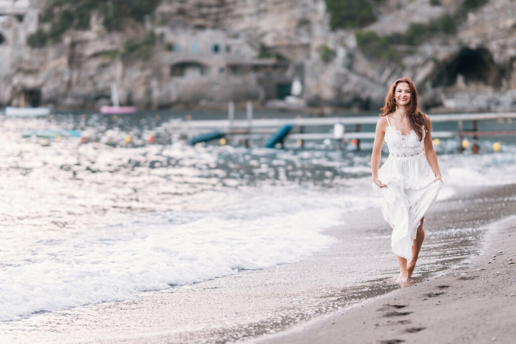pmphoto-38 Smiling woman in a white lace dress walking along the shoreline in Positano, Italy